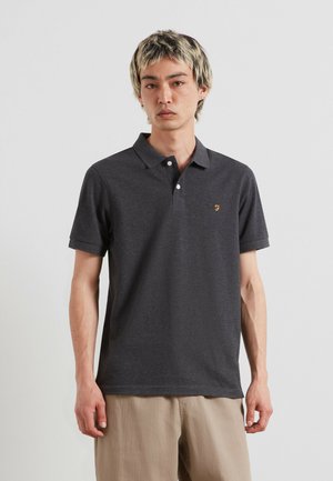 Giovane uomo con capelli biondi corti indossa una polo grigio scuro e pantaloni beige chiaro, in piedi contro uno sfondo chiaro uniforme.