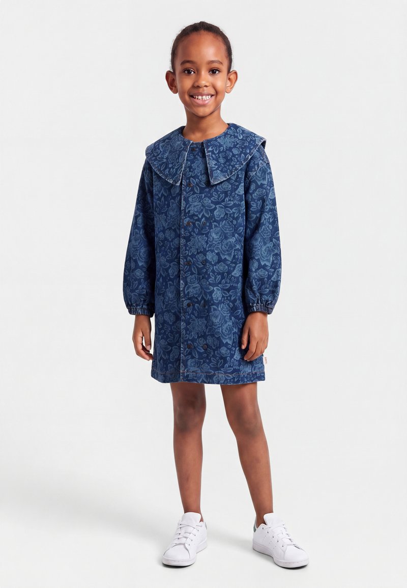 Jeune fille souriante, portant une robe bleue fleurie avec un grand col et des manches longues, associée à des baskets blanches, debout sur un fond uni.