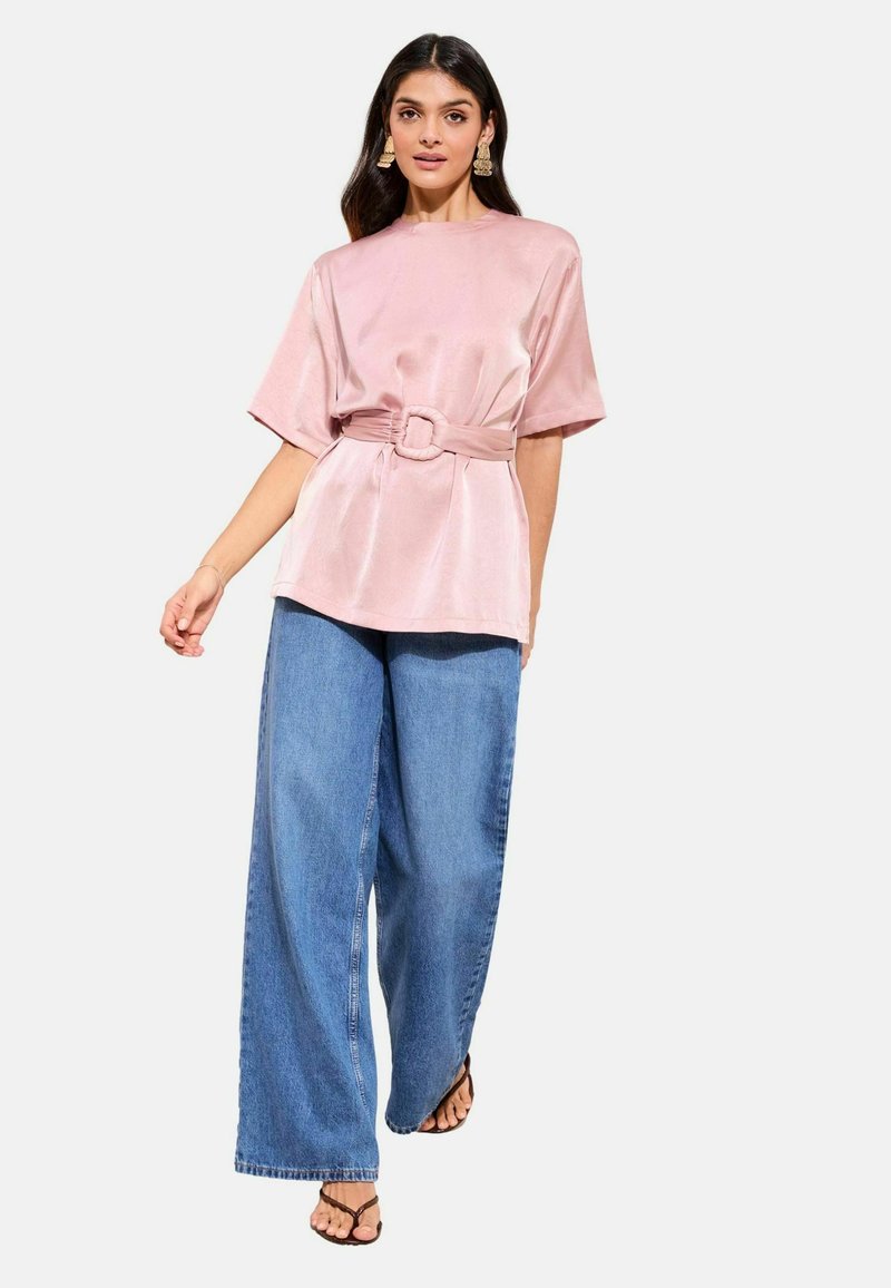 Sieviete, kas valkā rozā satīna topu ar jostu, plašām zilām džinsa biksēm un melnas sandales, stāv balta fona priekšā.