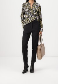 Blumenbluse in Schwarz, Gelb und Lavendel, kombiniert mit schwarzen Tailored Pants. Dazu tragen Sie schwarze Stiefel mit Absatz und eine beigefarbene Handtasche.