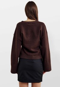 Brauner Strickpullover mit weiten Ärmeln und geripptem Saum, kombiniert mit einem schwarzen eng anliegenden Rock. Schlichtes Design, keine sichtbaren Muster oder Verzierungen.