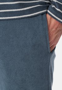 Tissu en éponge bleu avec une texture douce. Présente un motif rayé et une poche latérale, une coupe semi-ajustée, avec des détails de couture visibles.