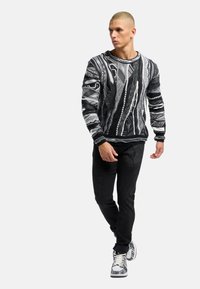 Pull en tricot à motifs noir et blanc, avec des rayures texturées et un col rond large, associé à un jean slim noir et des baskets à motifs.