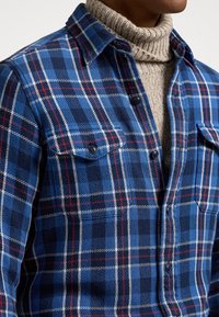 Camicia di flanella in blu, nero e bianco a quadri, con un design a bottoni e due tasche sul petto. Indossata sopra un maglione a collo alto color crema.