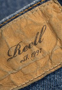 Etiqueta de cuero marrón con el texto en relieve "Reell est. 1997," cosida sobre una tela de mezclilla con hilos azules visibles y una superficie texturizada.