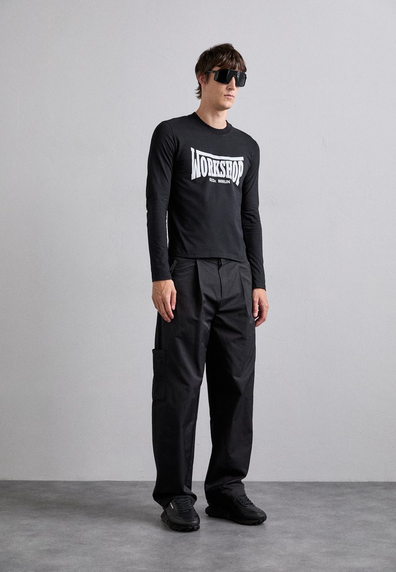 Schwarzes Langarmshirt mit weißen Grafiktext, kombiniert mit weiten schwarzen Cargo-Hosen mit Taschen. Das Modell trägt große schwarze Sonnenbrillen und Sneakers.