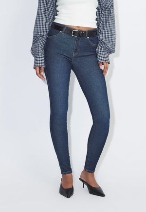 Mujer con vaqueros skinny oscuros, cinturón negro, top corto blanco, camisa de cuadros con mangas abullonadas y tacones negros de punta, de pie frente a fondo blanco.