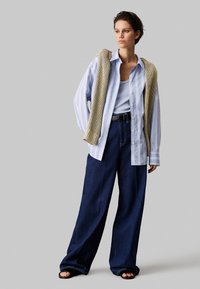 Lyseblå skjorte med knapper, parret med en ribbet tanktop, vide mørk denim jeans og et strikket beige sjal draperet over skuldrene.