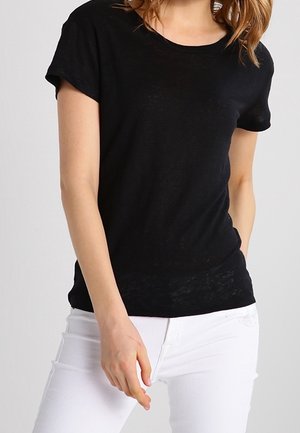 Kobieta w dopasowanej czarnej koszulce z krótkim rękawem i białych spodniach, stojąca na jednolitym jasnoszarym tle.