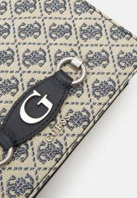 Bolso de tela beige con un diseño estampado en azul marino, que presenta un anillo de hardware redondo en color plata y una correa de cuero negro con un logo de metal en forma de 'G'.