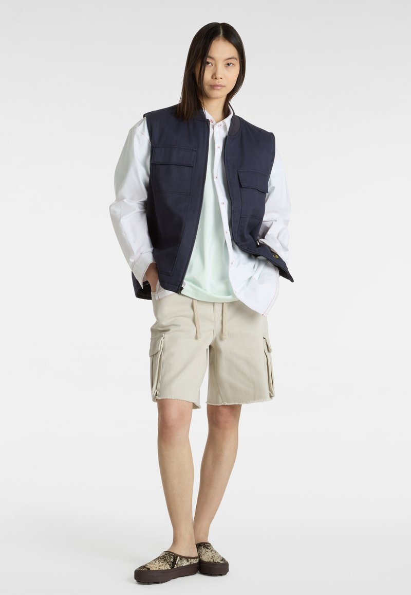 Chaleco sin mangas navy sobre una camisa blanca de botones, camiseta verde claro, pantalones cortos beige tipo cargo y zapatos tipo slipping con estampado.