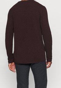 Maglione a maglia bordeaux con un motivo testurizzato, maniche lunghe, scollatura rotonda e orlo curvo, abbinato a pantaloni scuri.