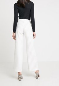 Valkoiset raidalliset leveälahkeiset housut korkealla vyötäröllä, yhdistettynä mustaan ribbikankaiseen pitkähiharaiseen puseroon ja hopeisiin käärmemuotoisiin korkokenkiin.