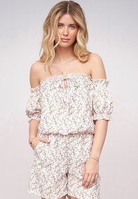 Blomstret off-shoulder romper i hvitt med rosa og grønne blomster. Puffermede ermer, volangdetalj ved halsen, nøkkelhullsdetalj og sidelommer.