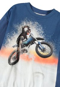 Kék, hosszú ujjú ing, amelyen egy motocross versenyző látható, amint akcióban van, alul pedig kékből narancssárgára gradálódik. Pamut kevert anyag.