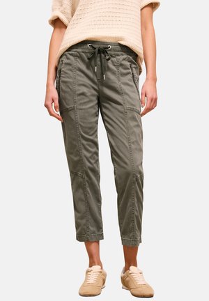 Person iført korte, olivengrønne bukser med snøre og lynlåslommer, beige striktop og beige sneakers, stående på en ensfarvet hvid baggrund.