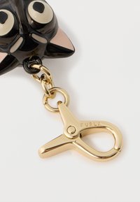 Musta muovinen avaimenperä kissadesignilla, jossa on suuret silmät ja vaaleanpunaiset korvat. Kiinnitetty kultainen metalliklipsi, jossa on kaiverrettu "FURLA". Suuri sileä pinta.