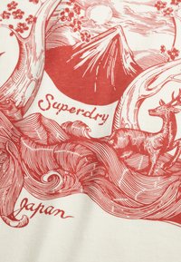 Biały materiał z czerwonym nadrukiem przedstawiającym góry, jelenia oraz stylizowane drzewa z kwiatami. Tekst zawiera słowa "Superdry" i "Japonia".