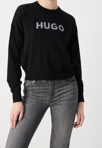 Schwarzer Strickpullover mit rechteckigem "HUGO"-Logo in Silber, kombiniert mit grauen Slim-Fit Jeans. Der Pullover hat gerippte Bündchen und einen gerippten Saum.