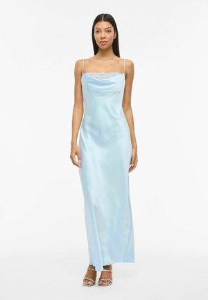 Frau mit langen dunklen Haaren, die ein hellblaues Satin-Nachthemd mit dünnen Trägern und Spitzenapplikation trägt, stehend vor weißem Hintergrund.