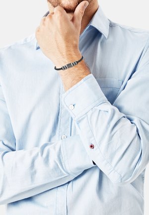 Braccialetto nero intrecciato presenta un accento metallico centrale con strisce orizzontali, indossato sopra una camicia a maniche lunghe azzurro chiaro.