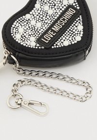 Μαύρο πορτοφόλι σε σχήμα καρδιάς με μεταλλικά τρουκς, ετικέτα "Love Moschino"· διαθέτει ασημένια αλυσίδα με κούμπωμα και φερμουάρ.