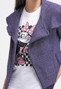 Chaqueta morada de cremallera con un tejido texturizado, que presenta un cuello amplio y un corte relajado, llevada sobre una camiseta blanca con un gráfico de un personaje de dibujos animados.