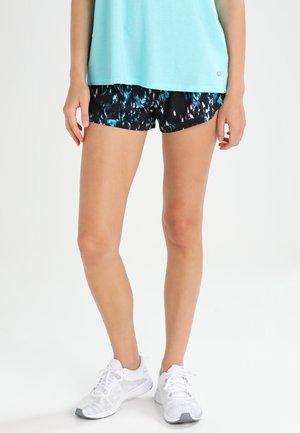 Femme portant un débardeur bleu, un short de sport noir à motifs et des chaussures de course blanches, debout dans une posture détendue.