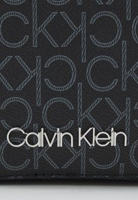 Materiale nero testurizzato con motivo ripetuto "CK" grigio; presenta il logo "Calvin Klein" argentato nella parte inferiore.