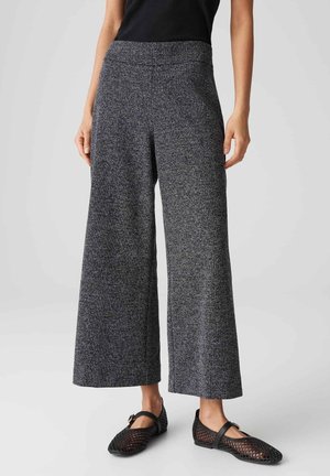 Vrouw draagt grijze broek met wijde pijpen en zwarte Mary Jane schoenen van gaas, staand tegen een effen lichte achtergrond.