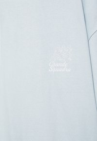 Tissu bleu clair avec un contour blanc de trois figures courant au-dessus du texte « Grande Squadra » sur la partie supérieure de la poitrine d'un vêtement.