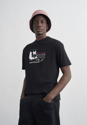 Mann iført rosa bøttehatt, svart T-skjorte med teksten "Jazz Café Fred Amsterdam City" og grafikk, og svarte kordfløyelsbukser med hendene i lommene.