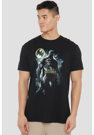 Mann trägt schwarzes T-Shirt mit großem Batman-Grafik und Fledermaussignal, steht vor schlichtem hellem Hintergrund.