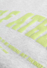 Felpa grigia con testo in rilievo di un vivace verde neon. La texture è morbida e leggermente pelosa, con una stampa a contrasto sul davanti.