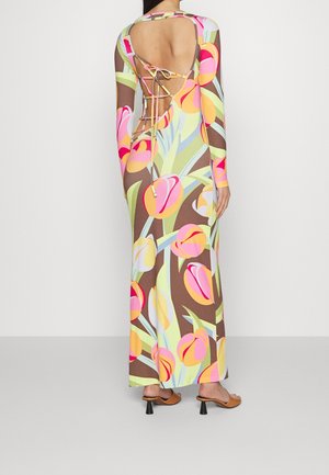 Kvinde iført en langærmet blomster maxi-kjole med åben ryg og snøre-detalje, kombineret med brune sandaler med hæl.