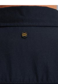 Tamno plavi materijal s malom crnom i zlatnom oznakom "PME Legend" ušivenom blizu gornjeg ruba.