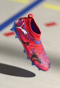 Botte de football synthétique rouge avec des accents bleus, surface texturée et motifs graphiques dynamiques en noir et bleu. Elle présente un design à tige basse et des crampons.