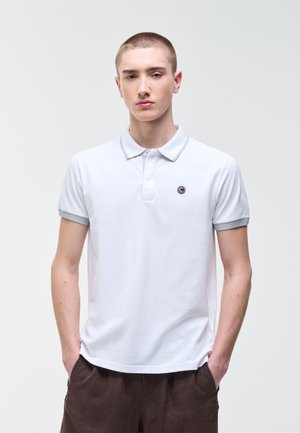 Joven con cabello corto, vestido con una camiseta polo blanca con ribete gris y pantalones marrones, de pie con las manos en los bolsillos contra un fondo liso.