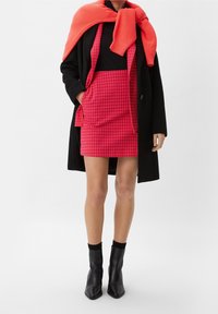 Czerwona spódnica w kratkę houndstooth z przednimi kieszeniami, w połączeniu z czarną bluzką i długim czarnym płaszczem. Jasnoczerwona dzianinowa sweterka przerzucona przez ramiona.
