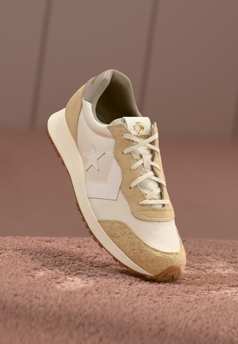 Adidași din suede și piele bej cu design în formă de stea albă, șireturi plate și talpă intermediară texturată. Talpa de cauciuc are modele de aderență.