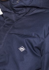 Marinblå vadderad jacka med hög krage, ribbad struktur och liten vit logotyp på nedre vänstra sidan. Inkluderar justerbar dragsko.