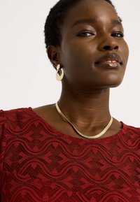 Collier doré au design courbé et à la finition texturée, assorti de boucles d'oreilles créoles, porté par un modèle vêtu d'un haut en dentelle rouge.