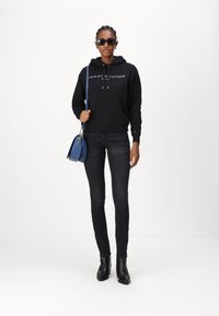 Sweat à capuche noir avec logo blanc, jean skinny foncé, bottines noires et sac à main bleu avec des accents en métal. Modèle portant des lunettes de soleil.