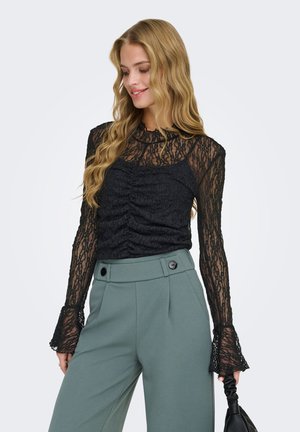 Femeie cu păr lung și blond, purtând un top negru din dantelă transparentă cu mâneci lungi și pantaloni verzi cu talie înaltă, ținând o geantă neagră.