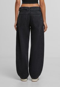 Henkilö, joka on pukeutunut korkeavyötäröisiin, leveälahkeisiin mustiin farkkuihin ja valkoisiin tennareihin, seisoo selkä kameraan päin yksinkertaista harmaata taustaa vasten.