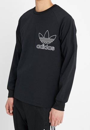 Man wearing black Adidas long-sleeve shirt with white trefoil logo and black pants with Adidas stripes.