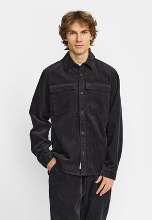 Jonge man met warrig haar, gekleed in een donkergrijs ribfluwelen overhemd met knopen en bijpassende broek, staand met één hand in de zak.
