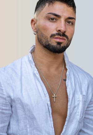 Man with a beard wearing a silver cross necklace, dressed in a light blue linen shirt with an open collar, featuring a smooth texture.