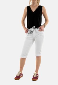 Zwarte mouwloze top met kantafwerking, witte cropped broek met opgerolde manchetten en een patroon riem, gecombineerd met rode sneakers.