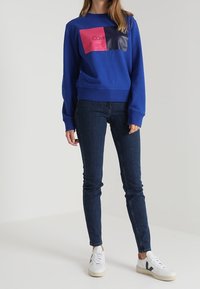 Modrá mikina se světlým designem a barevnými bloky, obsahující růžové a námořnické nášivky, spárovaná s tmavými džíny a bílými teniskami.